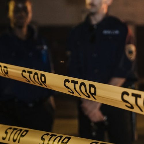 Two police officers standing behind a stop tape at night, scene partially illuminated by car headlights.