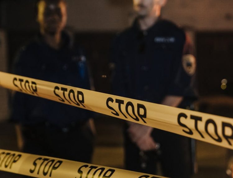 Two police officers standing behind a stop tape at night, scene partially illuminated by car headlights.