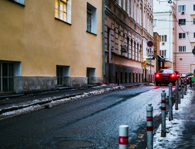 A deserted urban alleyway with cars, snow, and glowing windows at dusk.
