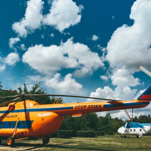 Two vintage helicopters on an airfield under a bright blue sky with clouds.