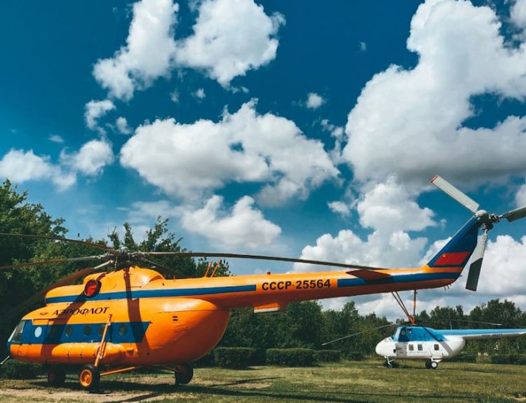 Two vintage helicopters on an airfield under a bright blue sky with clouds.
