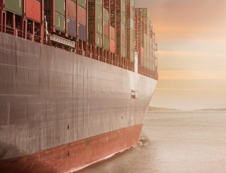 A colossal cargo ship loaded with containers navigates through calm waters against a vibrant sunset sky.