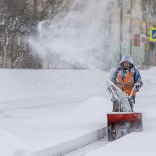Person using a snow blower to clear a city sidewalk during a heavy winter snowfall.