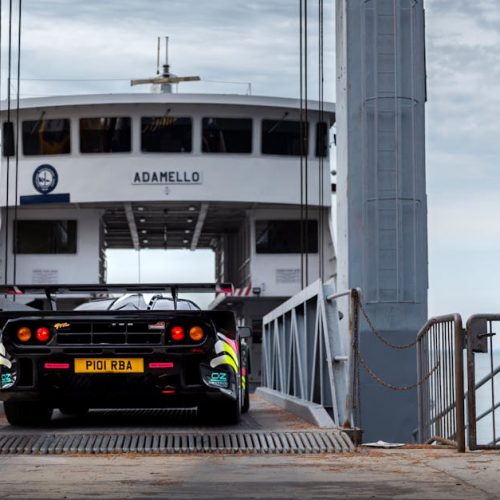A McLaren supercar driving onto a ferry named Adamello in the scenic Italian Alps.