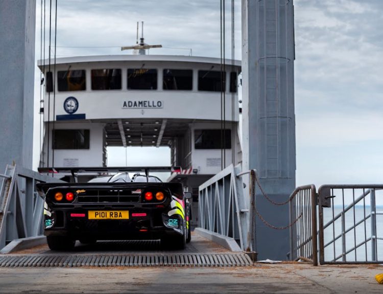 A McLaren supercar driving onto a ferry named Adamello in the scenic Italian Alps.