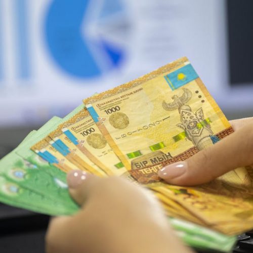 Hands holding Kazakhstan tenge bills with financial chart and graphs on computer screen.