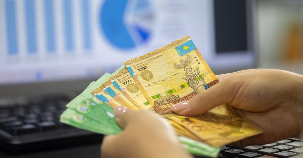 Hands holding Kazakhstan tenge bills with financial chart and graphs on computer screen.