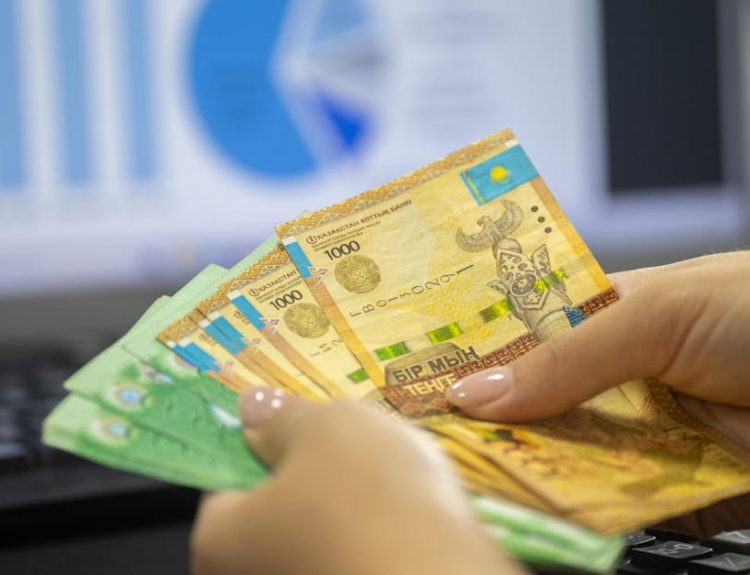 Hands holding Kazakhstan tenge bills with financial chart and graphs on computer screen.