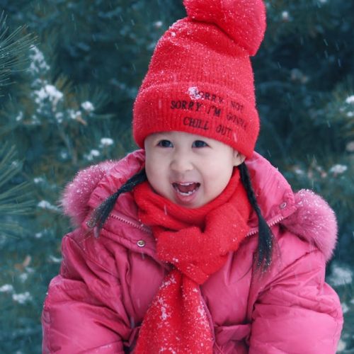 A young girl in winter attire smiles in a snowy outdoor setting, surrounded by pine trees.