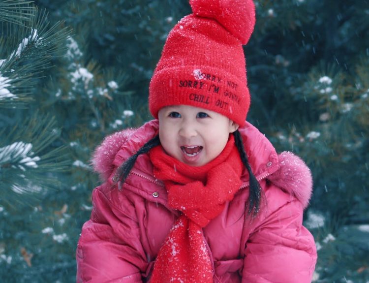 A young girl in winter attire smiles in a snowy outdoor setting, surrounded by pine trees.