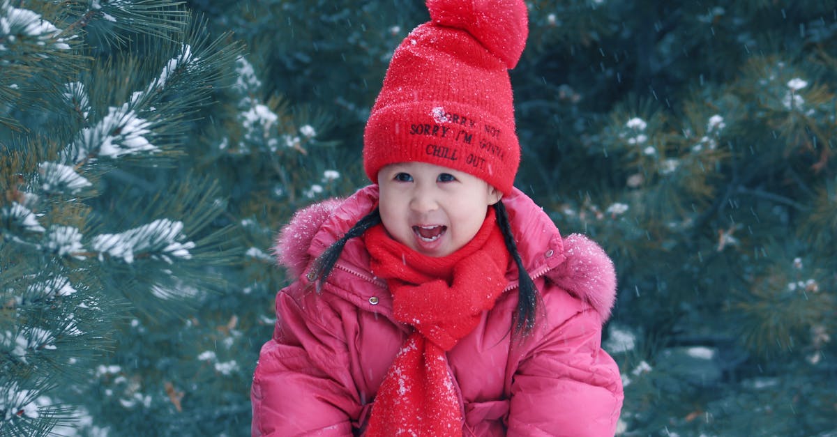 A young girl in winter attire smiles in a snowy outdoor setting, surrounded by pine trees.