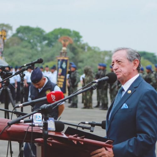 Side view of mature colonel with order standing near microphones on asphalt pavement behind army of soldiers near airplane monument under serene sky during national military holiday and looking away
