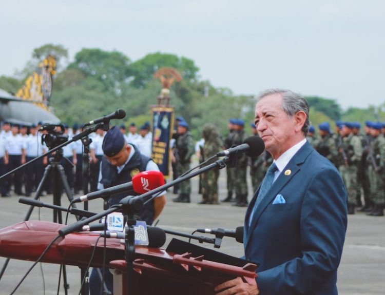 Side view of mature colonel with order standing near microphones on asphalt pavement behind army of soldiers near airplane monument under serene sky during national military holiday and looking away