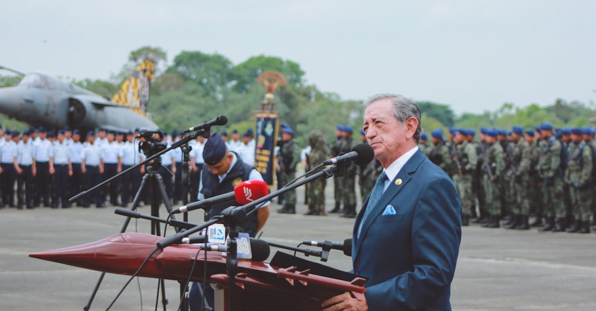 Side view of mature colonel with order standing near microphones on asphalt pavement behind army of soldiers near airplane monument under serene sky during national military holiday and looking away
