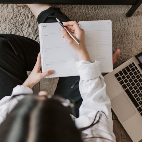 Top view of anonymous woman in casual wear sitting on floor with laptop and smartphone and creating plan on notebook while resting during break in modern living room