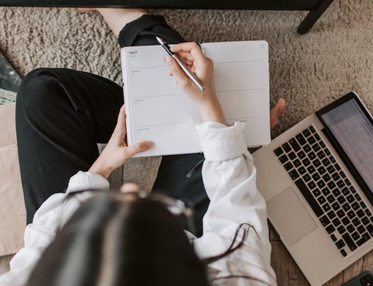 Top view of anonymous woman in casual wear sitting on floor with laptop and smartphone and creating plan on notebook while resting during break in modern living room