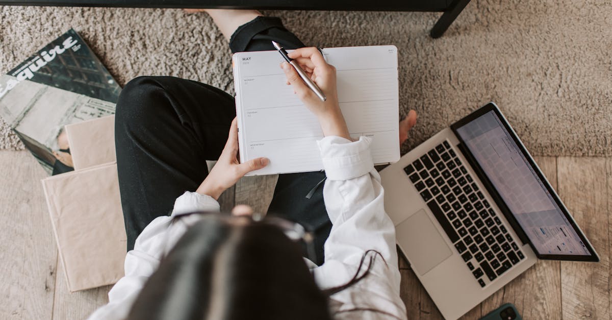 Top view of anonymous woman in casual wear sitting on floor with laptop and smartphone and creating plan on notebook while resting during break in modern living room