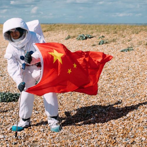 Person in a space suit holding a Chinese flag in a rocky desert setting, portraying an astronaut scene.