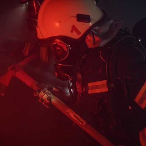 Firefighters in full gear with helmets and axes tackle a smoky emergency situation.