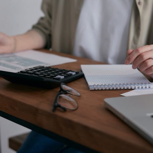 A woman manages finances at home, using a laptop and calculator on a wooden desk.