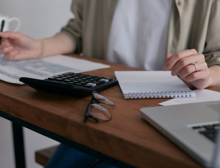 A woman manages finances at home, using a laptop and calculator on a wooden desk.