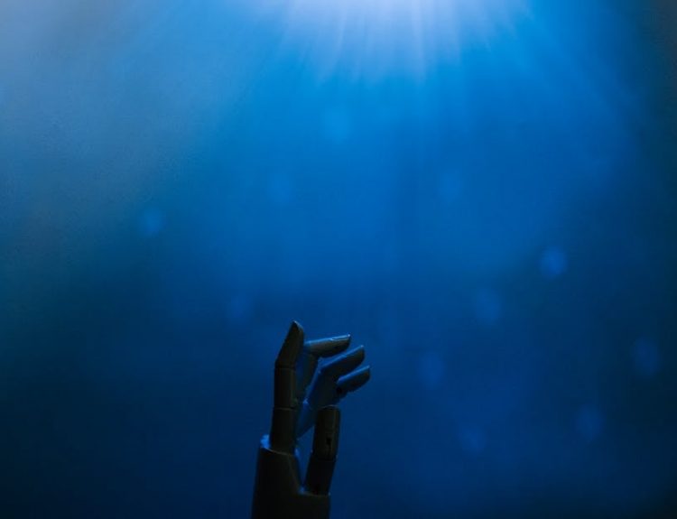 Silhouette of a robotic hand reaching towards glowing blue light in a futuristic setting.