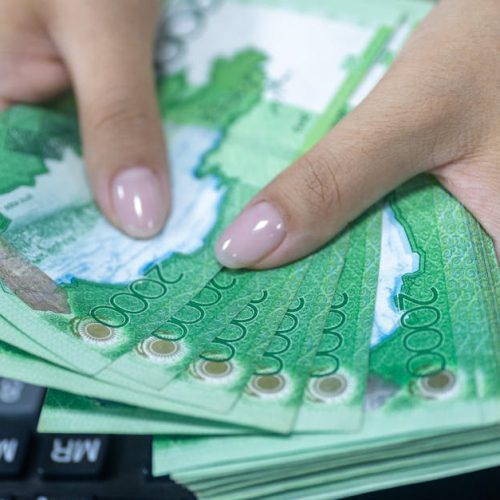 Close-up of hands counting Kazakhstani tenge bills beside a keyboard.