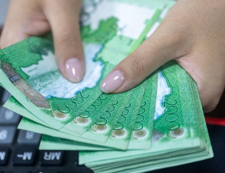 Close-up of hands counting Kazakhstani tenge bills beside a keyboard.