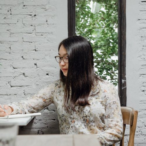 Pensive young Asian female writer in casual clothes and eyeglasses taking notes in diary while creating new article in cozy cafe