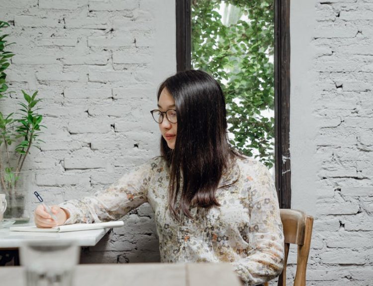 Pensive young Asian female writer in casual clothes and eyeglasses taking notes in diary while creating new article in cozy cafe