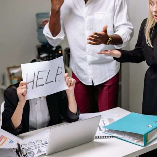 A group of employees in heated discussion, one holding a 'HELP' sign, indicates workplace stress.