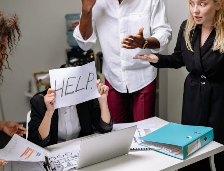 A group of employees in heated discussion, one holding a 'HELP' sign, indicates workplace stress.
