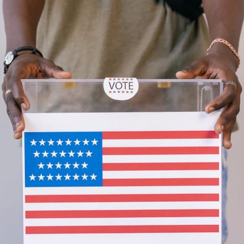 Close-up of a person placing a vote in a transparent ballot box with an American flag print.