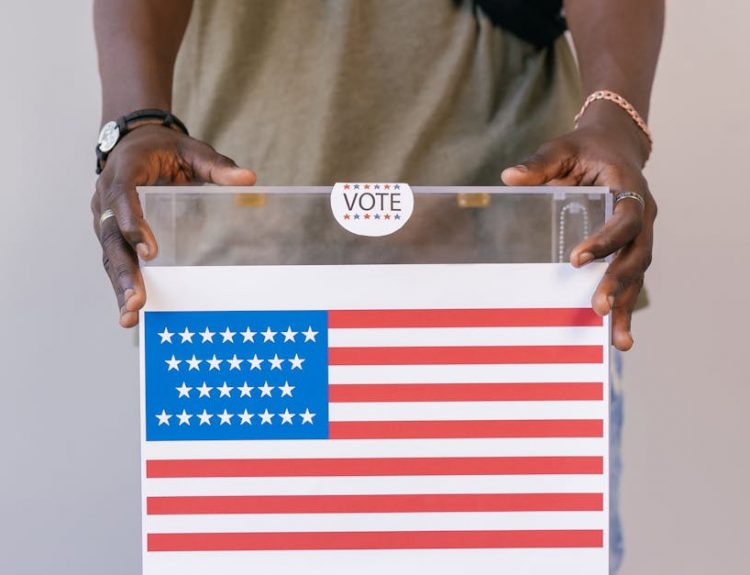 Close-up of a person placing a vote in a transparent ballot box with an American flag print.