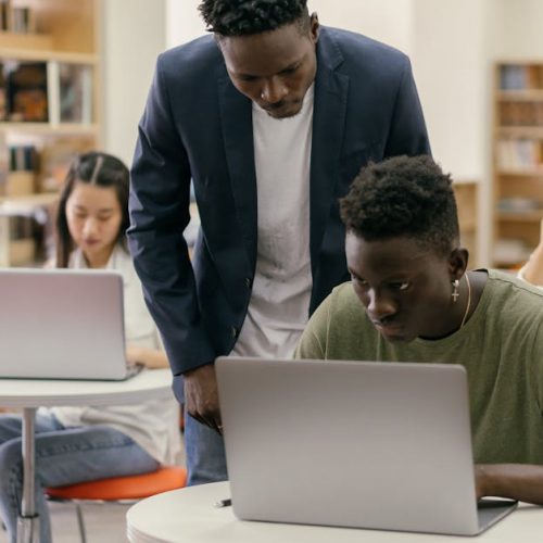 Students learning in a classroom setting with a teacher assisting and laptops on desks, creating an interactive education environment.