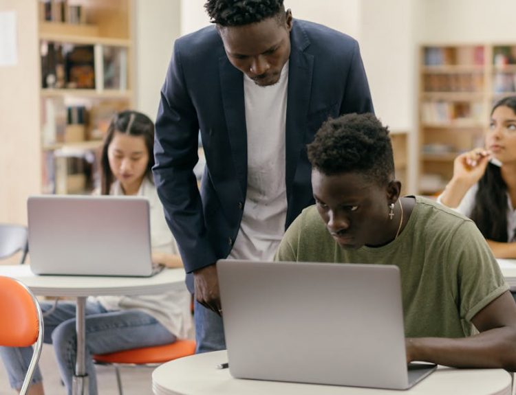 Students learning in a classroom setting with a teacher assisting and laptops on desks, creating an interactive education environment.