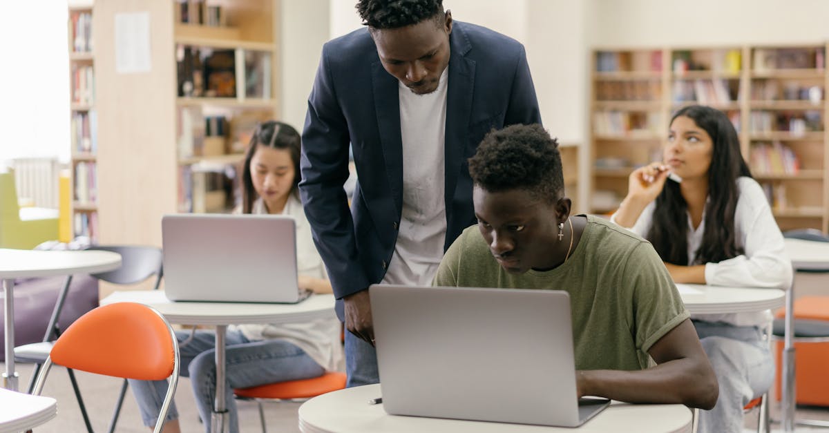 Students learning in a classroom setting with a teacher assisting and laptops on desks, creating an interactive education environment.