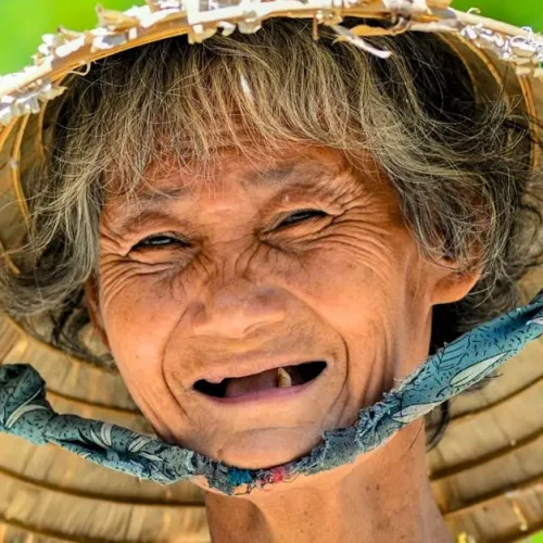 A vibrant portrait of a smiling elderly woman in a conical hat against a green background.