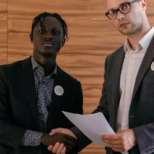 Two men in business attire shaking hands with agreement, promoting voting indoors.