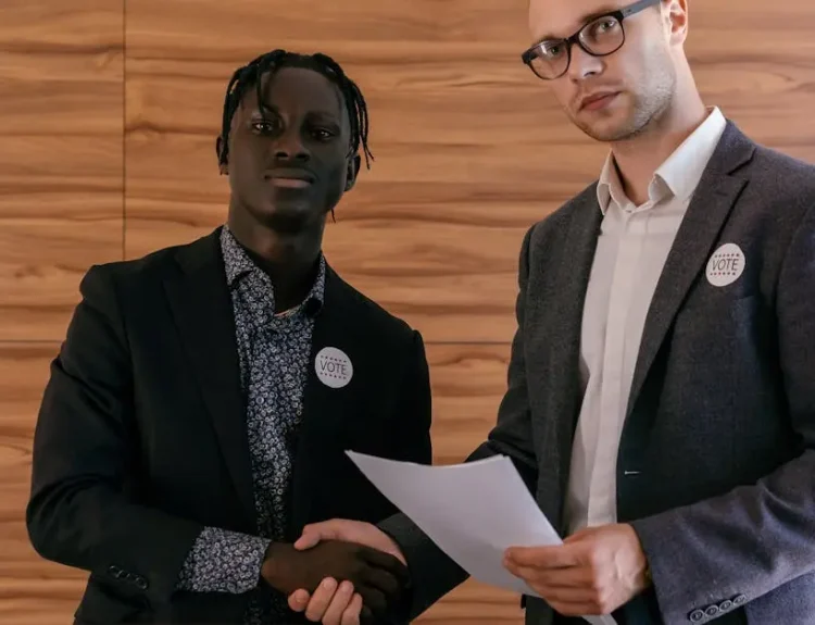 Two men in business attire shaking hands with agreement, promoting voting indoors.